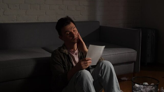 Man sitting on the floor in a dark living room, covering his face in despair after reading a upsetting letter, crumpled papers scattered around him, isolated and overwhelmed