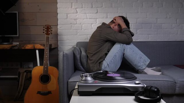 Man covering his face with hands, sitting on a sofa in a living room, experiencing mental health issues like depression, stress, and loneliness while listening to music from a vinyl player