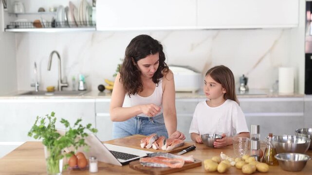 Mom and daughter are preparing festive salmon dish for family dinner. While cooking, mom peeks at laptop screen, clarifies recipe.