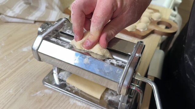 Hands feed fresh dough through a metal pasta machine on a floured wooden table, close view shows homemade cooking process, mood feels practical and focused