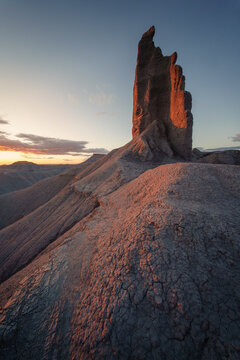 View of the rugged, arid landscape where a monolithic rock formation catches the warm glow of the setting sun painting the scene in orange and blue hues, Hanksville, Utah, United States.