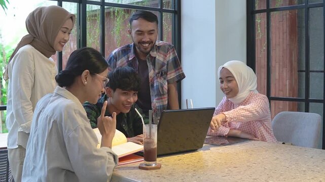 Asian friends gathered around laptop smiling as teenage student presents project to hijabwearing peers and mentor in bright cafe