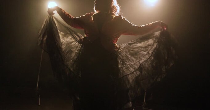 A female dancer raises her arms while lifting a dramatic flowing skirt on a dark smoky stage, silhouetted by bright theatrical backlights