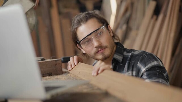 Professional male carpenter in safety glasses measuring a wood plank and checking online orders on a laptop in his workshop, focusing on precision and furniture manufacturing business