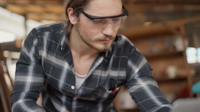Male carpenter in safety glasses working on a computer laptop in his woodshop, managing a small carpentry business and planning creative furniture projects