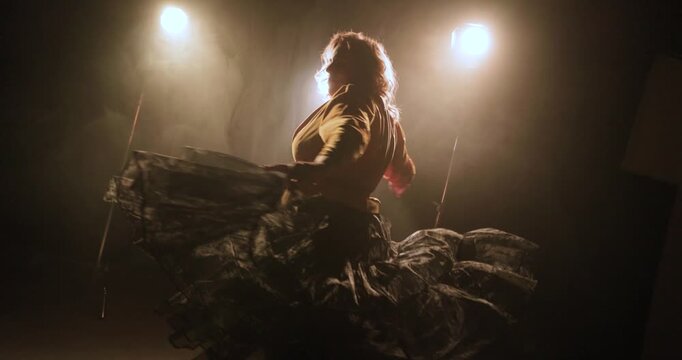 A female dancer spins on a smoky dark stage with arms outstretched, flowing skirt, and bright backlights creating a dramatic silhouette