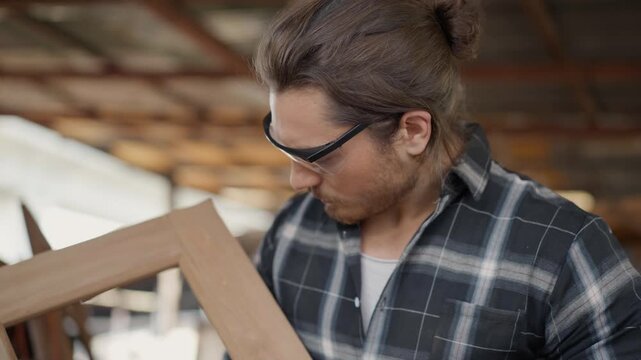 Professional carpenter inspecting a handmade wood frame in a carpentry workshop. Skilled craftsman wearing safety goggles, checking quality of timber project in a rustic industrial studio