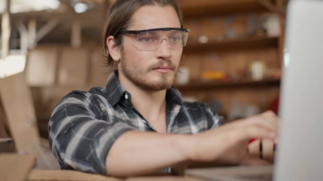 Focused male carpenter wearing safety goggles and using a pencil to mark wood in a carpentry workshop. Professional craftsman drafting a woodworking project in a rustic industrial studio