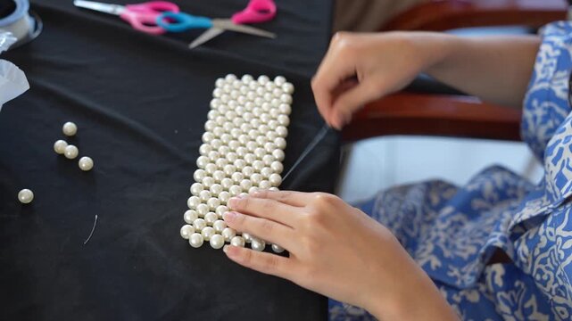 A medium high-angle shot of a designer working at a black studio table, meticulously weaving a structured grid of large white pearls.