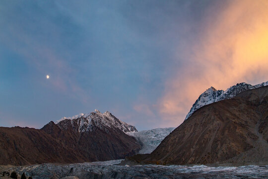 View of a majestic glacier winding through rugged, snow-capped mountains under a twilight sky with a crescent moon and warm, glowing clouds, Gilgit-Baltistan, Pakistan.