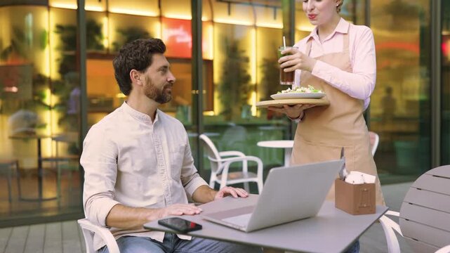 A waiter in an apron serves a salad and a drink to a man sitting at a table with a laptop in an outdoor cafe