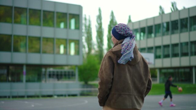 Woman in headscarf seen from behind standing on outdoor basketball court, candid moment observing surroundings, everyday life during cancer treatment