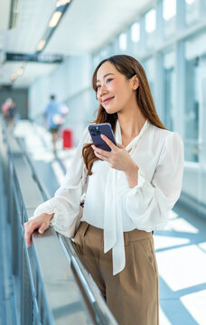 Asian Businesswoman standing with smartphone by railing in sunlit skybridge