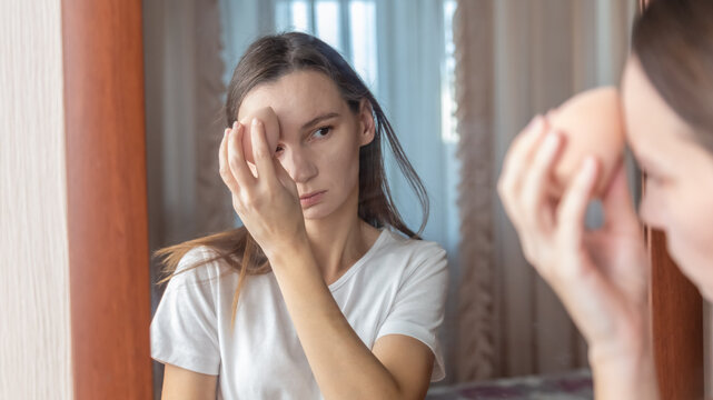 Young woman with problems skin applying cosmetic foundation with applicator near mirror indoors. Female using decorative makeup at home