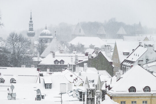 View of a snow-covered cityscape, roofs adorned with white blankets, church spires piercing the sky amidst a flurry of snowflakes, Tallinn, Estonia.