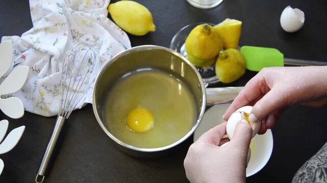 Close-up of hands cracking an egg into a bowl with visible yolk and egg white. Home cooking process with fresh ingredients and kitchen utensils on the table.