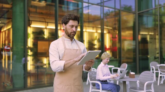 A focused male waiter wearing an apron uses a tablet device while standing outside a modern cafe with reflective glass windows