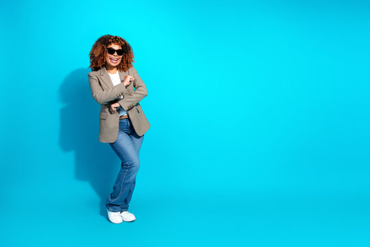 Young woman with curly hair wearing a blazer jeans and sunglasses smiles against a bright blue backdrop