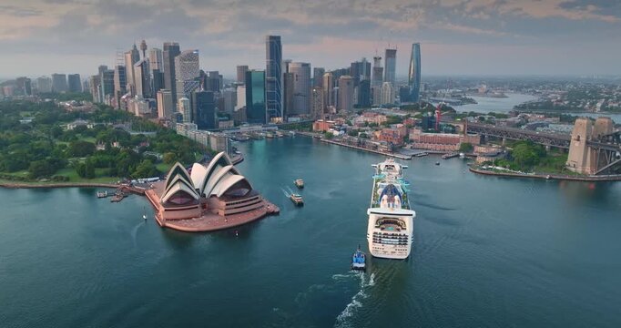 Sydney iconic Opera House and Circular Quay harbor featuring large cruise ship departing. Towering skyscrapers of the central business district under an early evening sky. Aerial drone flight panorama