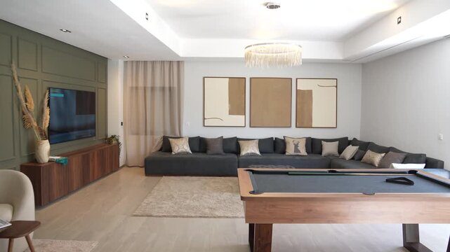 A smooth, bright cinematic pan of a spacious modern living area featuring a premium wooden pool table and a massive charcoal grey sectional sofa. The room is styled with a minimalist neutral palette.