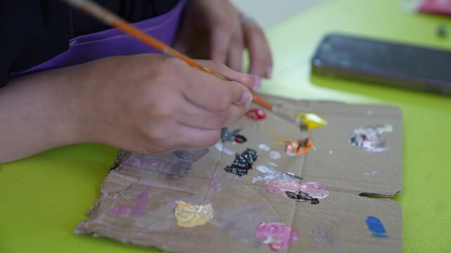 A detailed close-up shot of a person&rsquo;s hands using a fine-tip paintbrush to mix black and orange paint on a makeshift cardboard palette. The video captures the artistic process on a vibrant green surf
