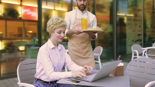 A smiling waiter in an apron serves a fresh salad to a woman sitting at an outdoor cafe table with a laptop and drink