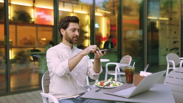 A man with a beard sits at an outdoor cafe table, holding his phone to take a picture of his salad and drink while a laptop is open nearby