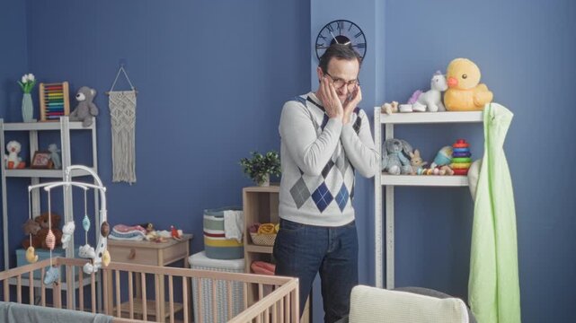 Man with hands clasped near face by wooden crib and hanging mobile in building nursery among shelves and toys; fatherhood bonding tenderness.