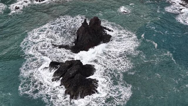 Drone aerial view of ocean waves crashing on volcanic rocks, dramatic seascape.
Aerial drone shot of powerful ocean waves crashing against dark volcanic rocks, creating white foam patterns in a dramat