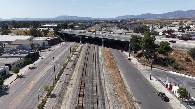 Cinematic daytime aerial drone footage captured hovering above train tracks in Los Angeles, facing toward a freeway bridge where vehicles speed past overhead.