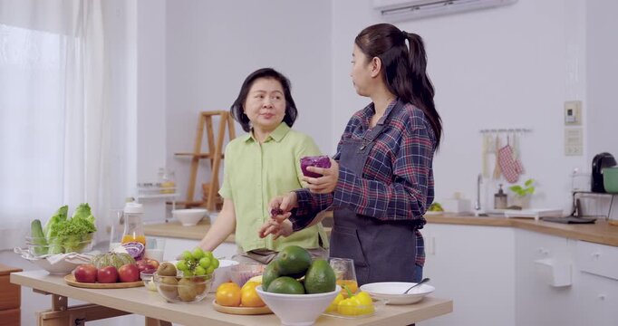 Asian senior mother talking with adult daughter holding purple cabbage in kitchen, family cooking preparation, communication, bonding, wellness, retirement lifestyle, teamwork, healthy food, organic