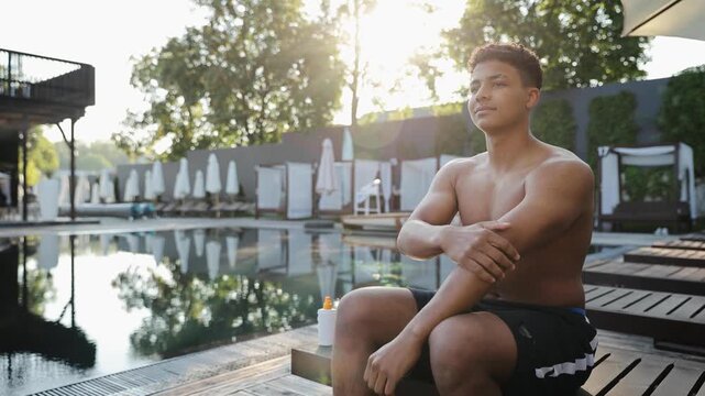 Young man sitting by pool, applying sunscreen to his shoulder. Practicing skincare and sun protection during summer