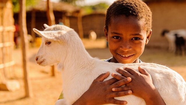 African child holding small white goat kid, close-up portrait with gentle smile looking at camera in rural village setting
