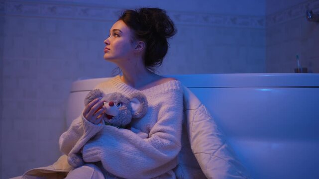 Sad young caucasian woman with depression sitting on the bathroom floor at night, hugging a stuffed toy. She eventually finds a moment of peace