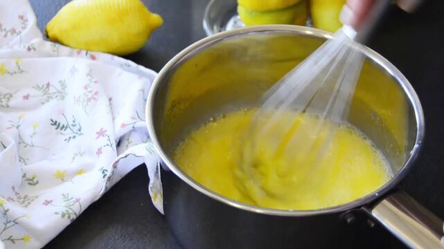Close-up of hands whisking lemon curd in a saucepan with smooth creamy texture forming. Homemade dessert preparation with fresh lemons and baking process.