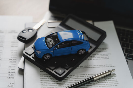 Blue toy car model on calculator with financial document showing numbers and key, pen and laptop on desk, representing car finance price calculation, auto loan value assessment, vehicle cost estimatio
