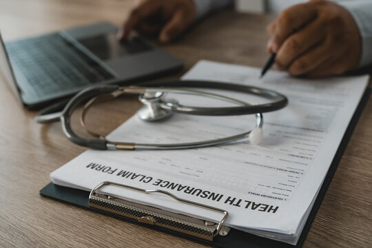 Medical insurance claim filing and healthcare policy concept with stethoscope on form clipboard, hand writing details with pen and laptop on wooden desk for health document paperwork process.