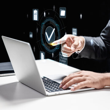 Businessman using a laptop while interacting with a digital holographic interface featuring a checkmark, symbolizing data verification, security, quality assurance, and project success