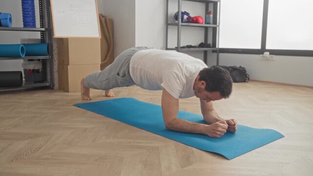 Man holding plank on forearms on blue yoga mat inside gym building with shelving, ropes and kettlebells visible; focus and endurance.