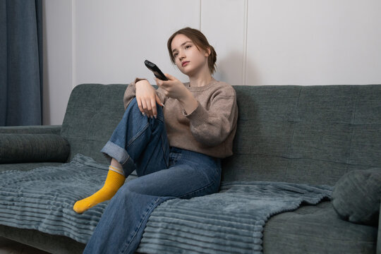 Young girl sitting on a comfortable couch at home, feeling bored and apathetic while navigating channels with a tv remote control, highlighting screen time and leisure indoors