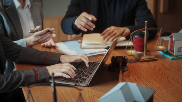Business meeting at wooden table showing hands, laptop, legal gavel and scales of justice, documents and model house, warm lighting