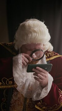 Vertical shot of Caucasian nobleman wearing wig and rococo embroidered coat using smartphone with magnifier indoors