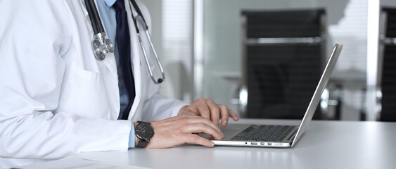 Fototapeta premium Medical professional typing on laptop, reviewing patient data, consulting online, using telehealth technology. Medicine, healthcare and science concept