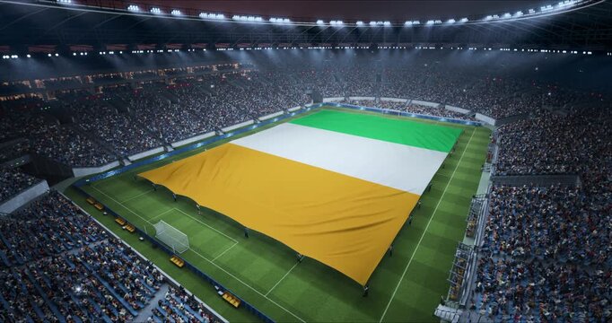 Professional Stadium With Soccer Game Championship With Flag of C&ocirc;te d'Ivoire Spread Across Pitch Before Football Match Start. Cheering Crowds of Fans Surround the Field. Aerial Static Wide Shot