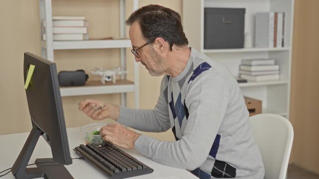 Man holds fork to mouth eating salad at computer in building, hands on bowl and keyboard, wearing glasses and argyle sweater while seated at desk; concentration.