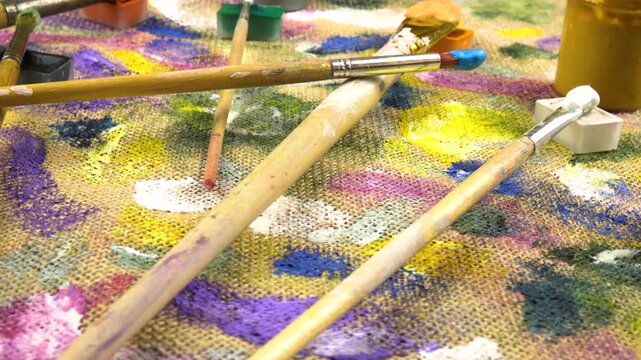 Jars with multi-colored paints, brushes are laid out on a beige burlap cloth with spots of different shapes and colors from gouache and acrylic paints, background, side view