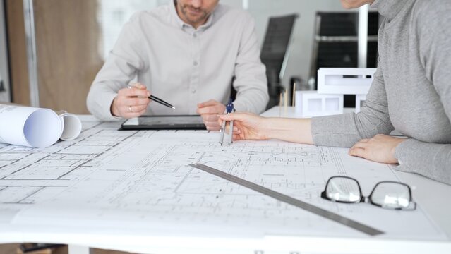 Architects are teamwork planning a building design project, reviewing detailed blueprints with a compass and a ruler, working on technical drawings and a digital tablet at the office, close up