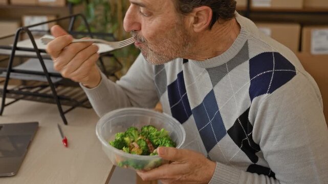 Man holding fork to mouth eating salad amid cardboard boxes and parcels at a small business office building; focused break.
