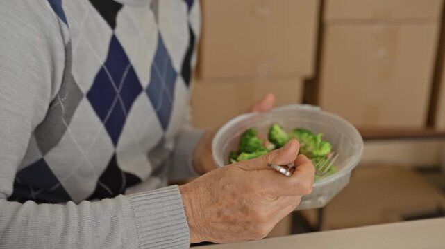 Man eating salad from plastic container, holds fork to mouth beside cardboard boxes in building; contentment small business.