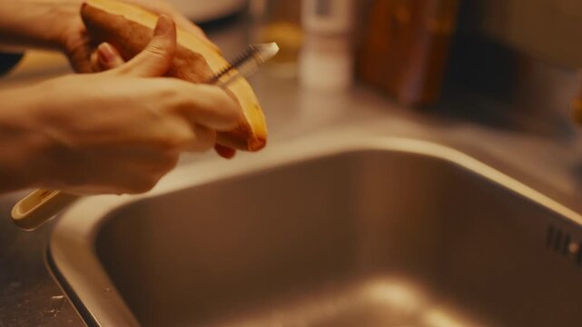 Close-up shot of female hands peeling raw sweet potato with a peeler over a kitchen sink. Warm lighting, selective focus with blurred metal countertop and wooden cutting board in the background.